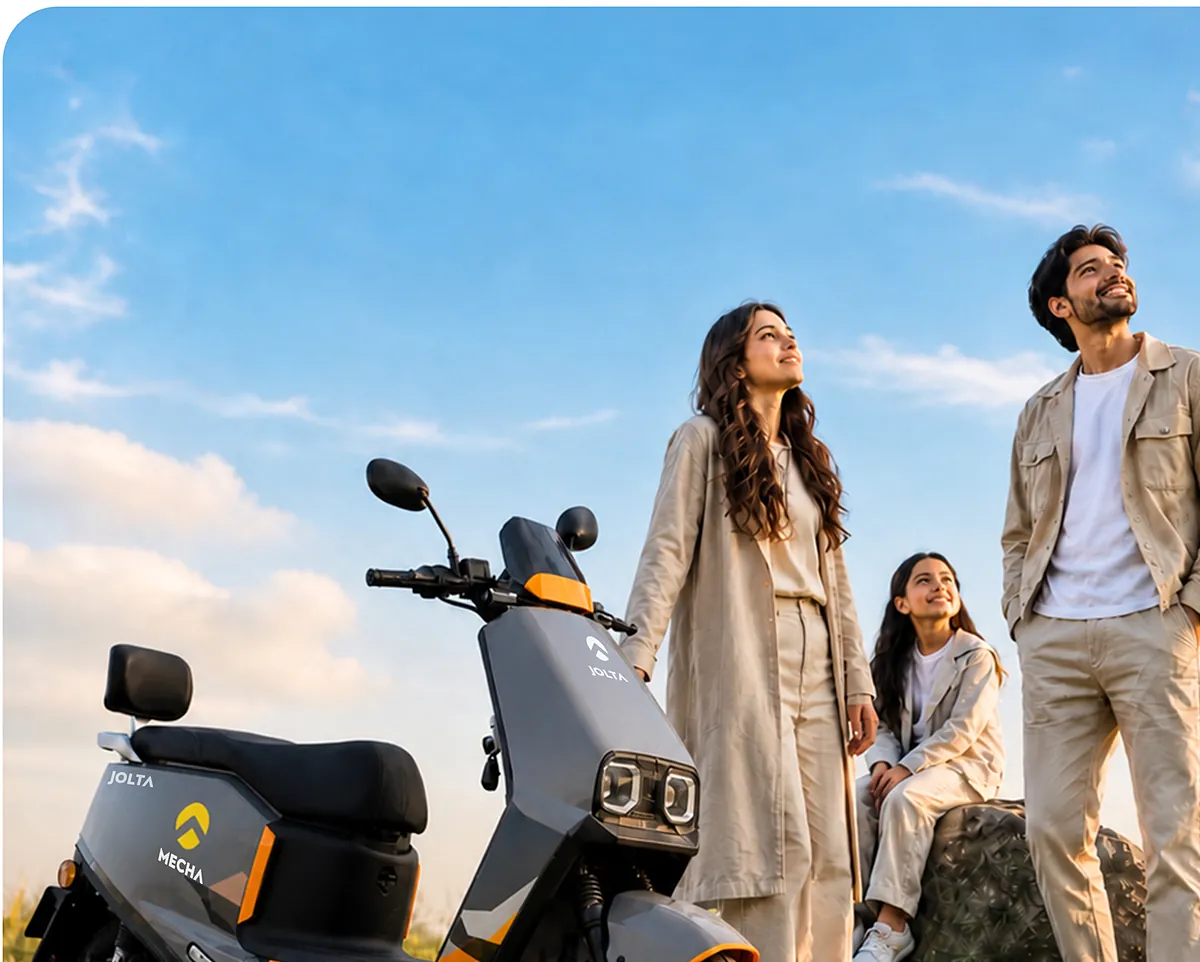 A family stands on a grassy hill next to a Jolta Mecha electric scooty
