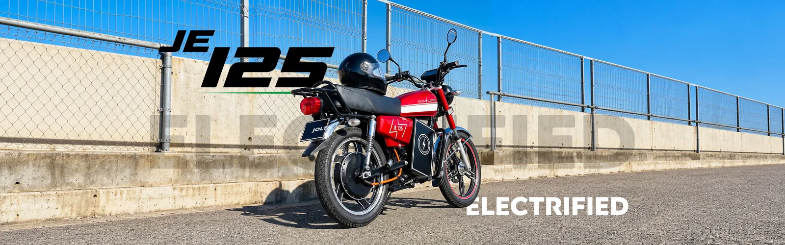 Red Jolta JE-125L electric motorcycle parked by a fence under a blue sky
